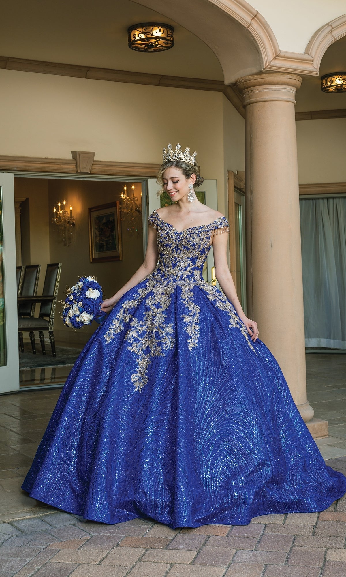 A woman in the Quinceanera Dress 1636 By Dancing Queen—sparkling royal blue with gold embroidery—wears a crown and holds a bouquet while standing before a columned building.